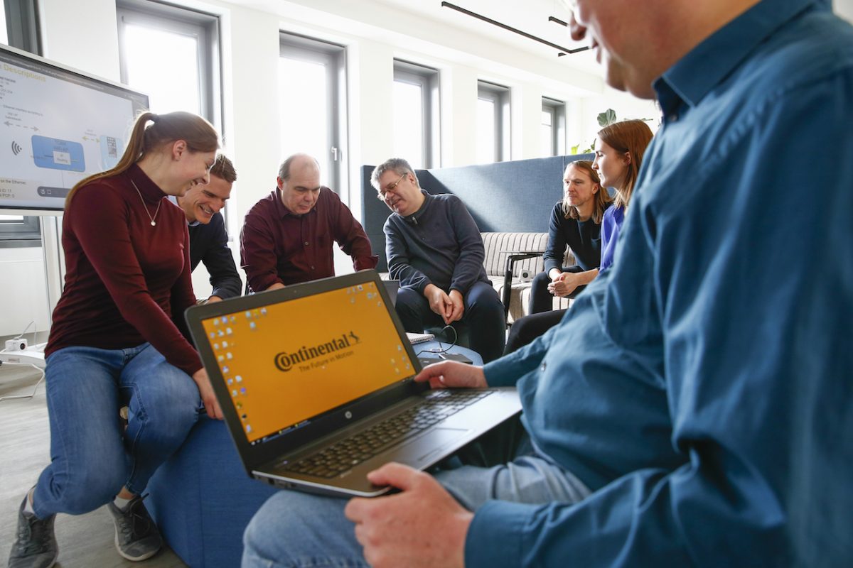 People sitting around each other. A man holds a laptop in the foreground